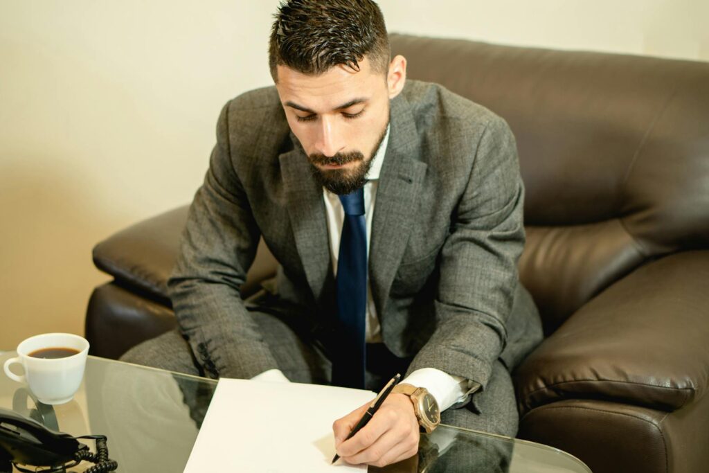 A businessman wearing a suit signs papers at a desk with focused attention.