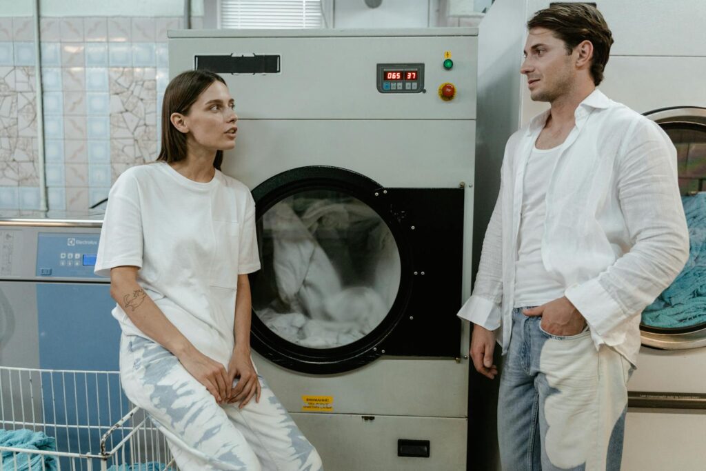 Two adults in a modern launderette having a casual conversation by washing machines.